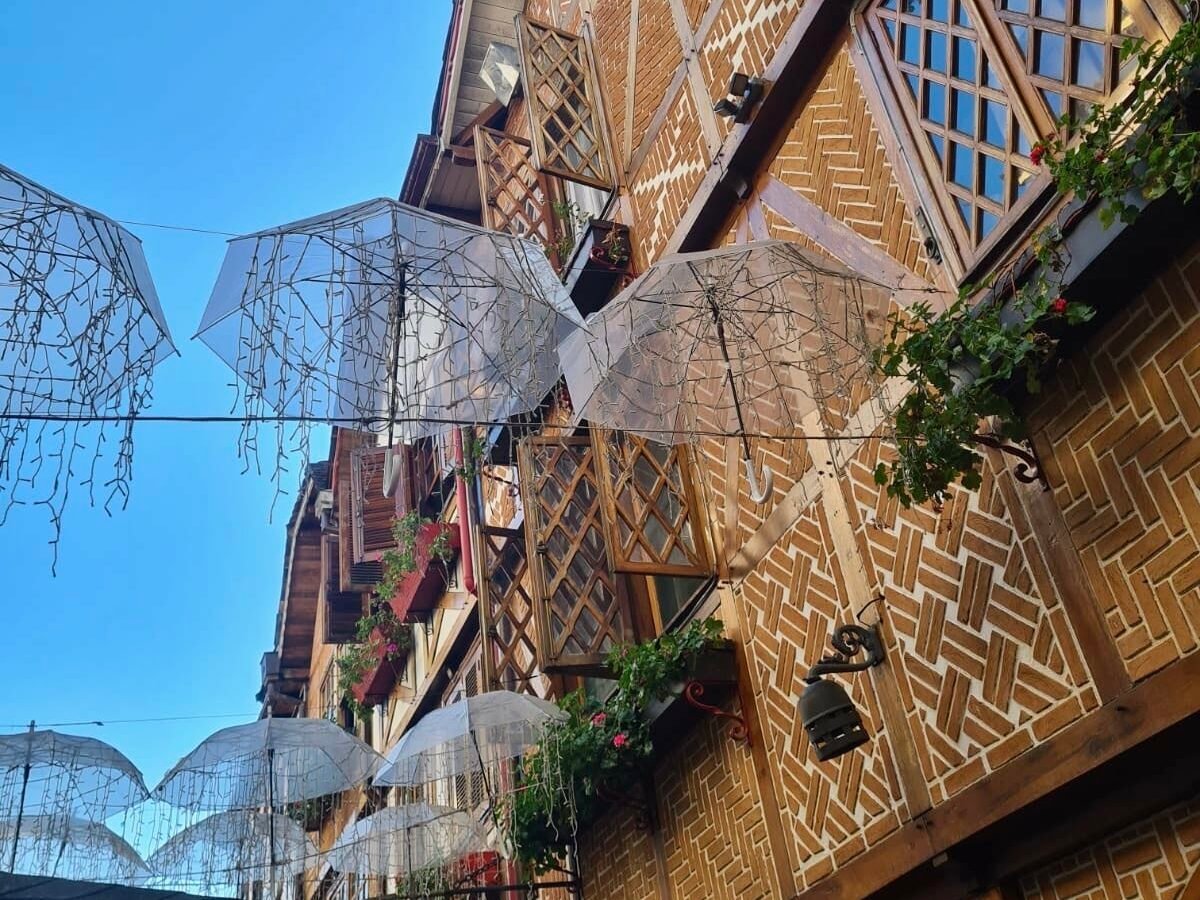 Rua entre os prédios com arquitetura suíça na Vila Capivari, onde há guarda-chuvas com piscas-piscas pendurados no ar. O céu está azul. Representa o post sobre o que fazer em Campos do Jordão.