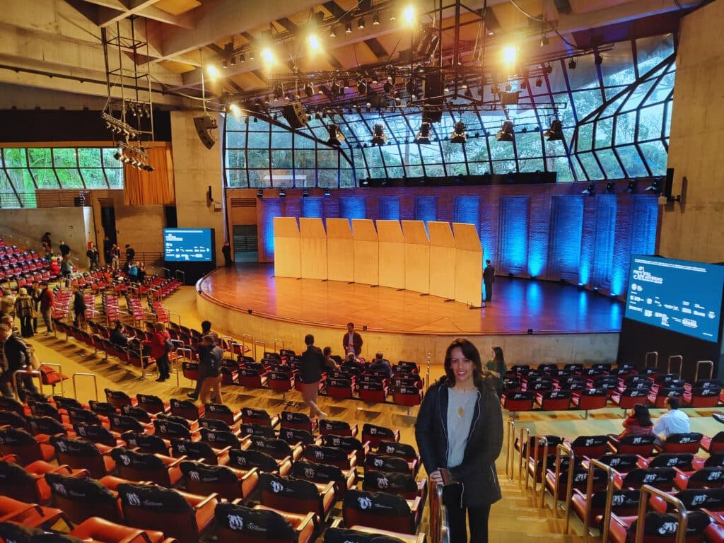 Foto do interior do Auditório Claudio Santoro em Campos do Jordão. Há vários assentos pelo ambiente. Uma mulher posa e sorri para a foto, se apoiando em um dos assentos no centro da imagem. Ao fundo está o palco com diversas iluminações e dois telões que o ladeiam. Há janelas no topo da parede.