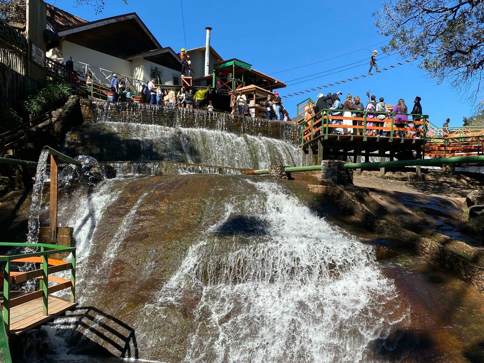 A imagem mostra uma cachoeira da Ducha de Prata em Campos do Jordão. Há pontes sobre a ducha onde as pessoas caminham. No topo se vê uma casa e uma ponte de tirolesa. O céu está azul.