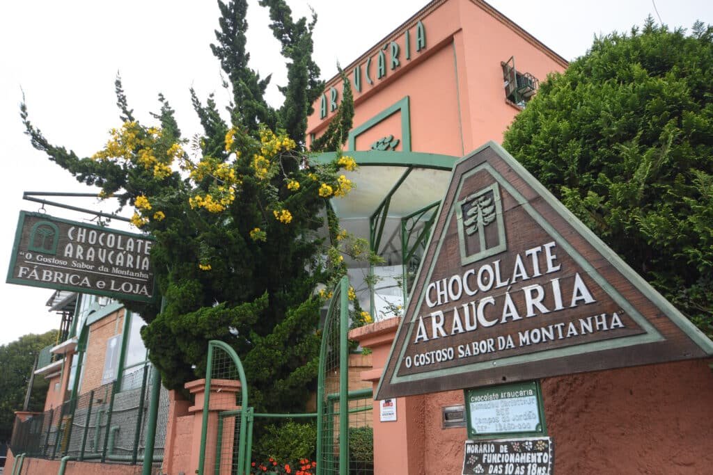 Fachada do prédio da fábrica de Chocolate Araucária em Campos do Jordão. Na direita há uma placa triangular de madeira indicando o nome e logo do lugar. No fundo, à esquerda, há outra placa igual, porém retangular. Há árvores e flores ao redor do prédio. O portão principal é verde e de ferro. O céu está nublado.