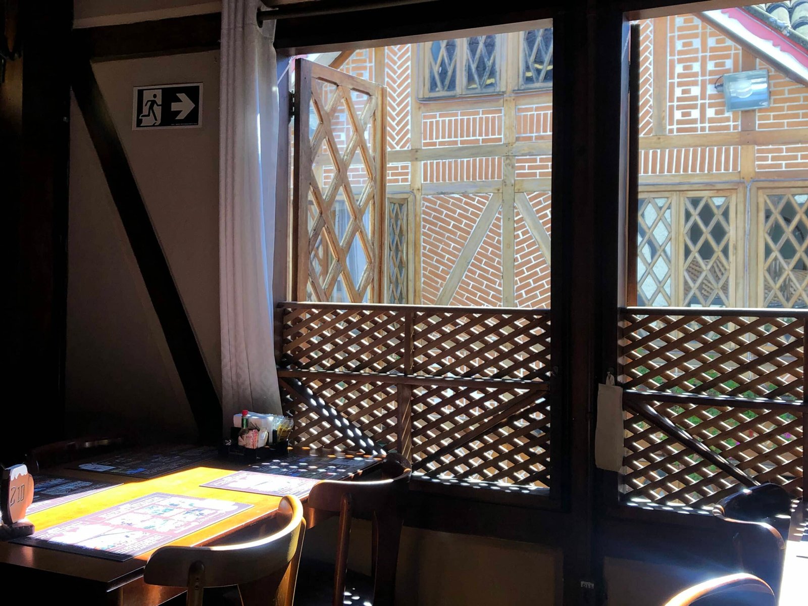 A foto mostra um pouco do interior da Cervejara Baden Baden. A decoração é majoritariamente feita por móveis de madeira, prezando pela arquitetura suíça. Vemos uma mesa com quatro lugares ao lado de uma janela no segundo andar do estabelecimento. A vista é para o prédio do lado.