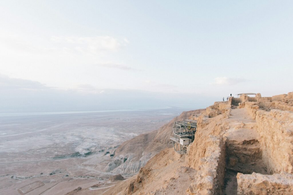 Ruínas de pedra em um penhasco com vista para uma paisagem desértica do Masada National Park, com algumas pessoas em pé perto de uma grade sob um céu claro. A foto faz parte do post de eSIM Israel.