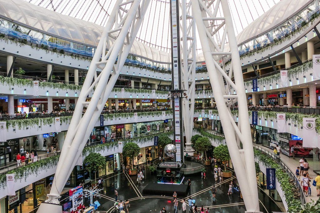 Interior do Khan Shatyr Entertainment Center, de vários andares com grandes vigas de aço brancas, lojas, área verde e pessoas caminhando. Imagem para ilustrar post sobre eSIM Cazaquistão.
