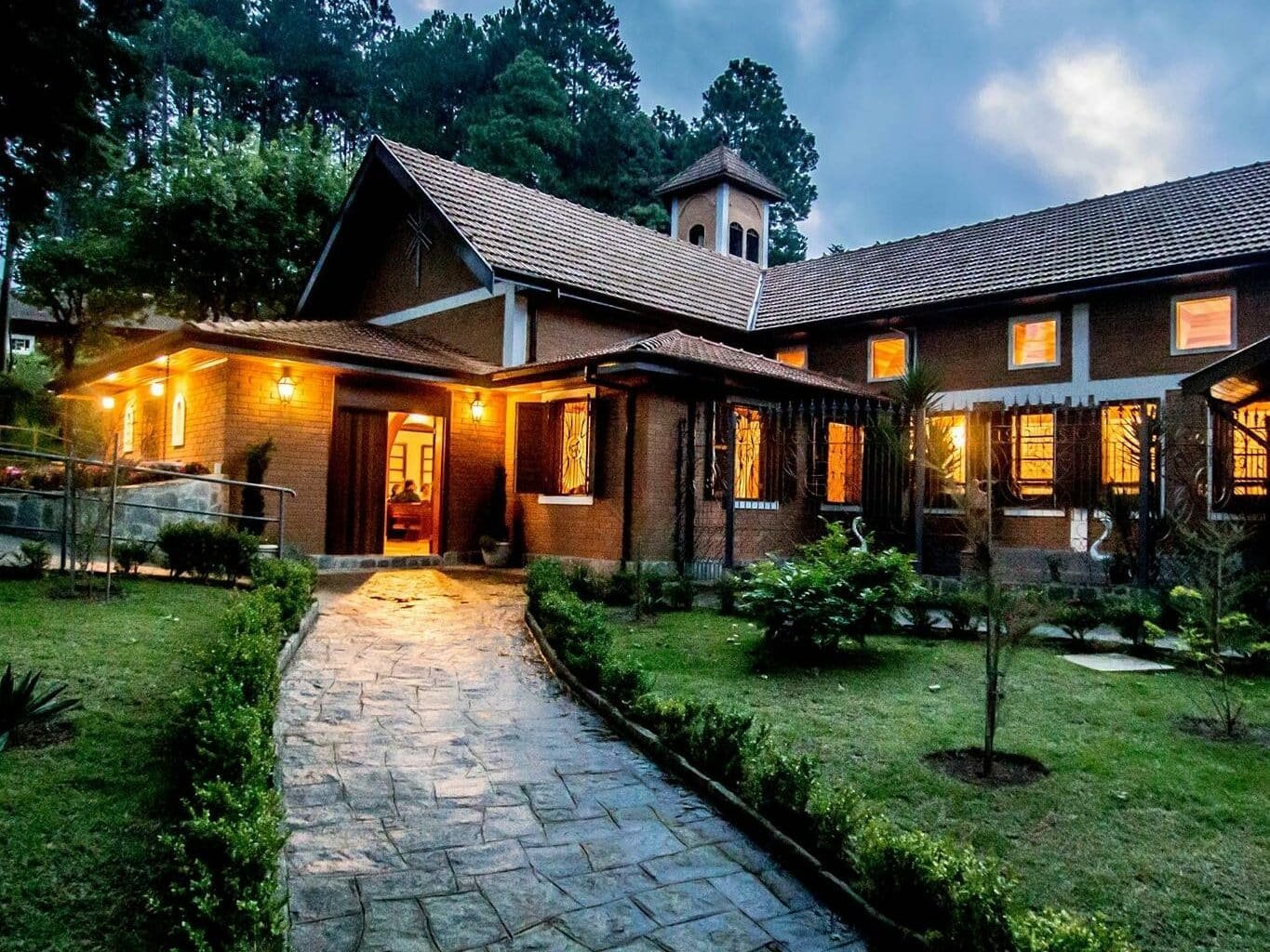 Foto tirada do Mosteiro de São João, em Campos do Jordão. Mostra um caminho de pedras entre um jardim com grama verde, que leva até a porta do mosteiro. É uma construção feita de tijolos, com vários cômodos e janelas. As luzes estão acesas. O céu está azul, porém está quase anoitecendo. Há árvores atrás do mosteiro. A foto ilustra o post de o que fazer em Campos do Jordão.