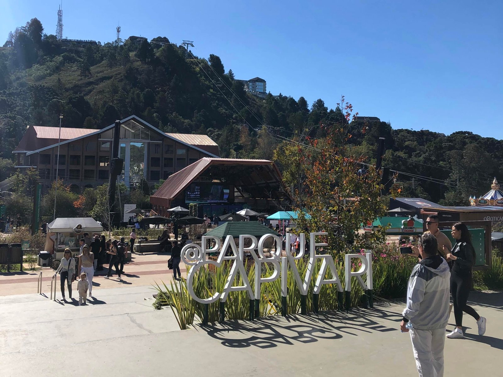 Entrada do Parque Capivari, em Campos do Jordão. No calçadão do parque já vemos um letreiro no chão com o nome do parque entre alguns arbustos, ideal para tirar fotos. No fundo vemos um palco, barraquinhas de comida, pessoas caminhando e um monte por onde sobe um teleférico. Representa o post sobre o que fazer em Campos do Jordão.