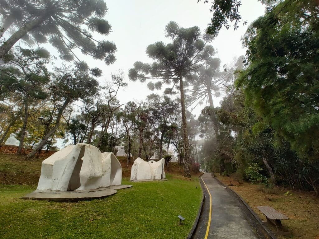 Foto de uma área ao ar livre no Parque Museu Felícia Leirner, em Campos do Jordão. Há uma leve neblina no bosque. Algumas esculturas brancas estão sobre o gramado à esquerda. Há um caminho pavimentado à esquerda.
