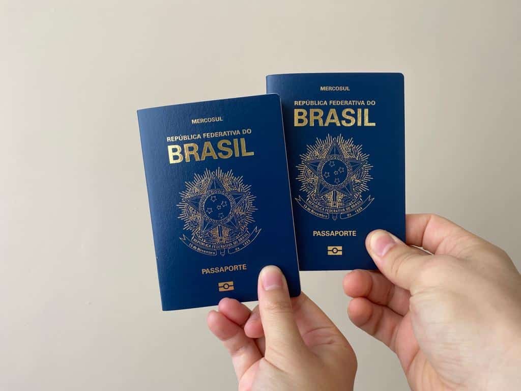 Mão feminina à direita, e mão masculina à esquerda, cada uma segurando um passaporte brasileiro fechado, em frente a um fundo bege