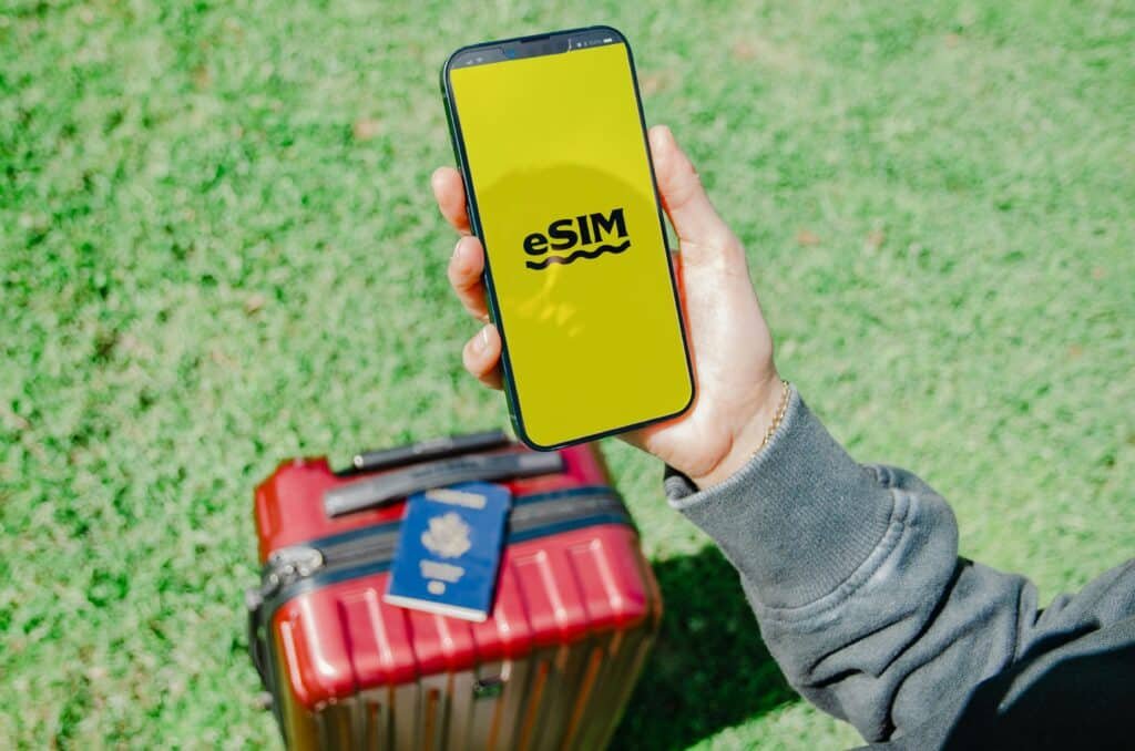 Uma pessoa segura um smartphone com a inscrição "eSIM" em uma tela amarela, acima de uma mala vermelha e um passaporte, na grama verde. Ilustra o eSIM Xangai