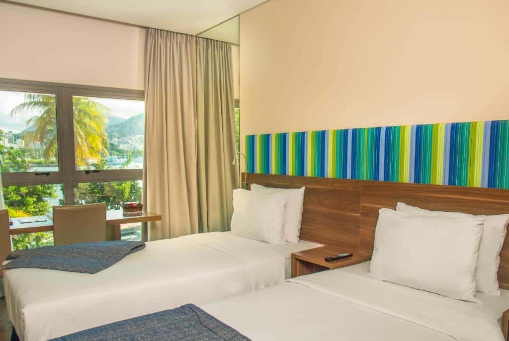 quarto do Prodigy Santos Dumont com duas camas de solteiro, cabeceira única e uma mesa de casal no meio das camas, além de mesa de trabalho ao fundo, em frente da janela e com cortina blecaute