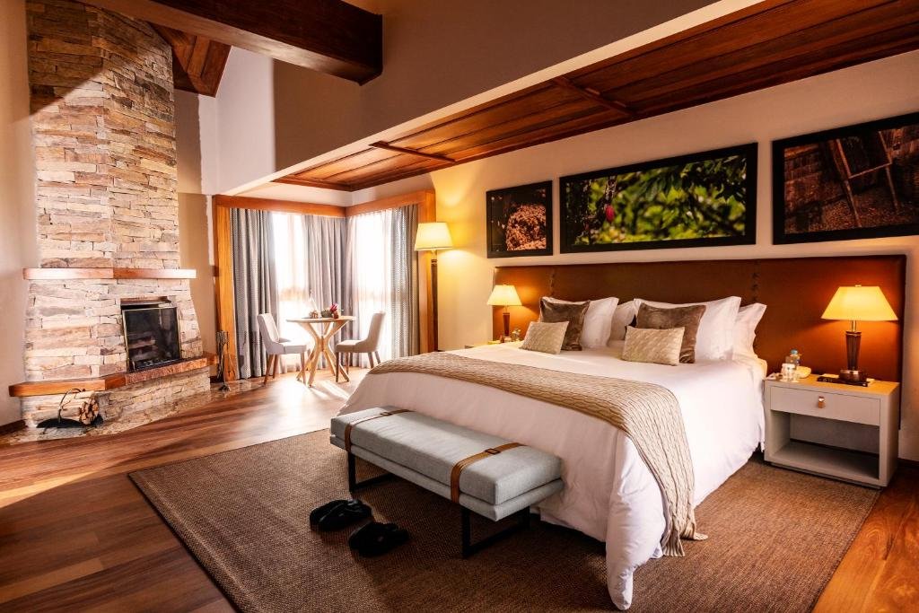 Quarto no Bendito Cacao Resort e Spa em Campos do Jordão. O ambiente é luxuoso, com uma cama de casal no centro sendo ladeada por duas pequenas cômodas com abajur em cima. Atrás da cama há uma parede com três quadros. Do lado esquerdo da cam há uma lareira e uma mesa com duas cadeiras na frente de uma janela.