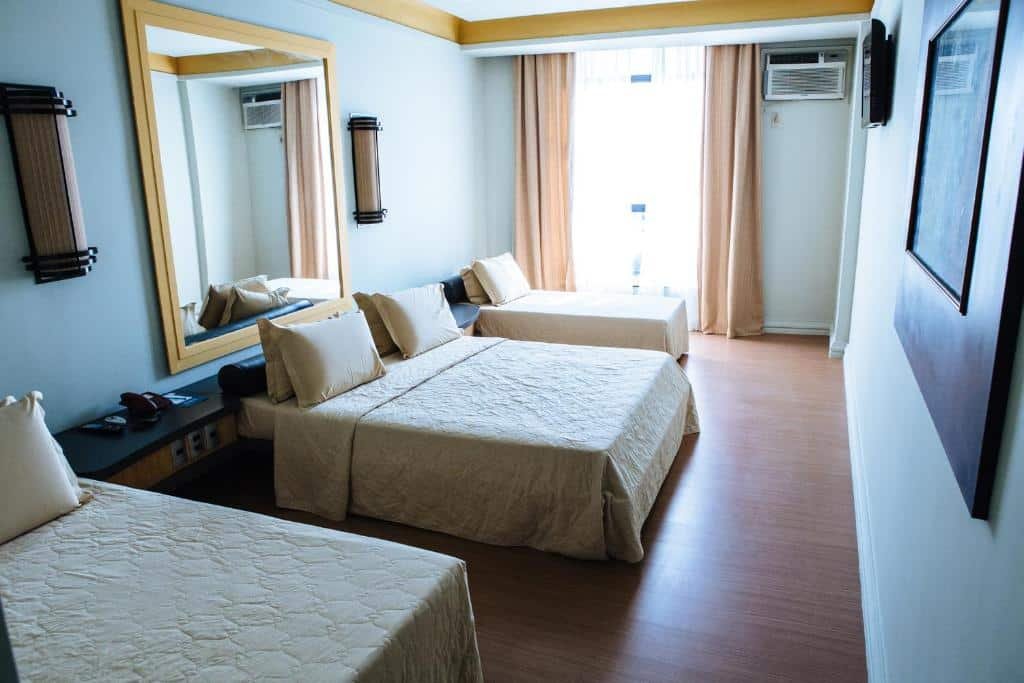quarto do Diamond Hotel, com uma cama de casal ao centro e duas camas de solteiro em cada lado, além de TV em frente à cama de casal, e ar condicionado ao fundo