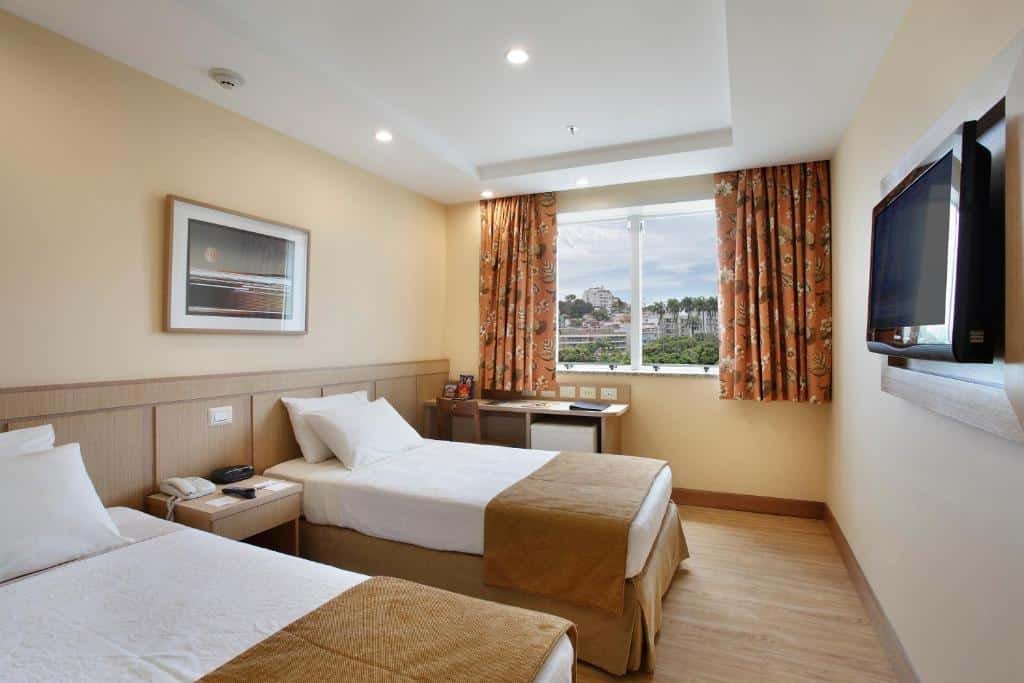 quarto do Windsor Flórida com duas camas de solteiro, cortina blecause na janela, mesa de trabalho abaixo dela, e TV na parede em frente à cama