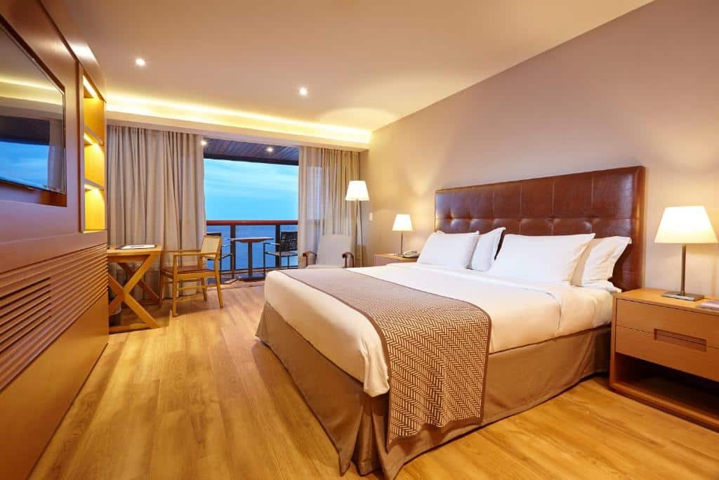 suíte do portobay, com TV plana em frente à cama de casal extensa, mesa de cabeceira com abajures, mesinha de trabalho e sacada ampla, com vista para o mar