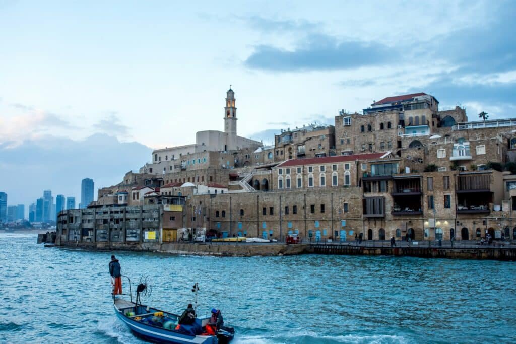 Um pequeno barco na água perto dos antigos edifícios de pedra de Jaffa, com o horizonte da cidade ao fundo sob um céu nublado. A foto faz parte do post de eSIM Tel Aviv.