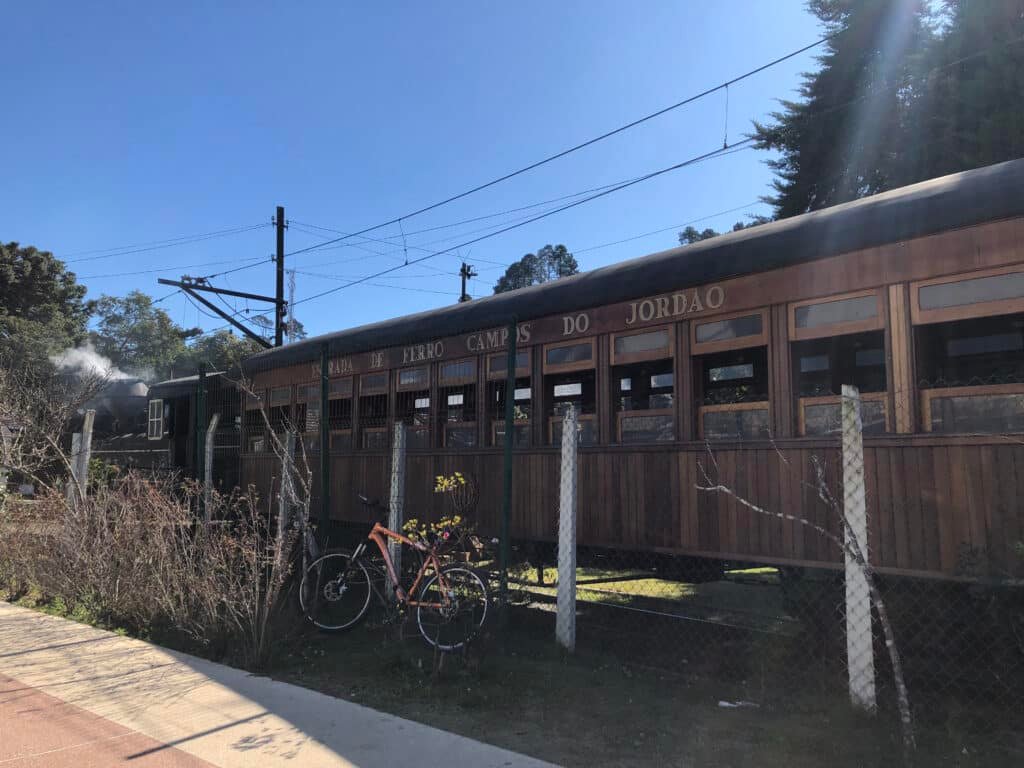 Foto do trem Maria Fumaça em Campos do Jordão. A parte frontal do trem por onde sai a fumaça está no canto esquerdo da foto, enquanto o vagão de madeira com janelas toma conta do centro da foto. Há um jardim e uma grade de ferro à sua frente. O céu está azul e sem nuvens.  